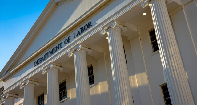 The image shows the facade of the U.S. Department of Labor building, featuring tall white columns in a classical architectural style. Sunlight casts long shadows across the structure, highlighting the fluted detailing and the bold black lettering of the b