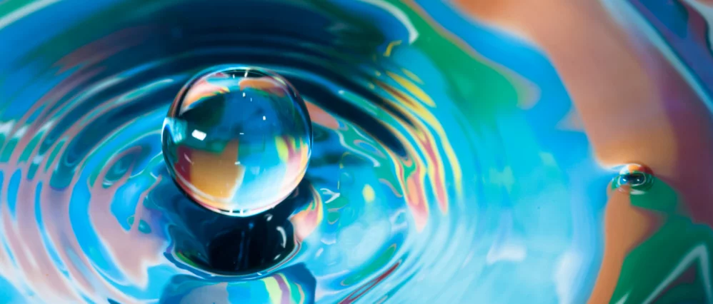 Close up of a clear water droplet suspended above rippling water with vibrant blue, green, and warm tones reflecting across its surface. The droplet resembles a miniature globe, with swirling colors that evoke the appearance of Earth.