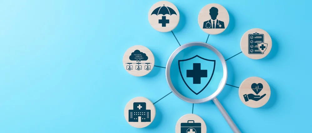 Magnifying glass centered on a shield with a medical cross icon, surrounded by connected wooden discs displaying healthcare related symbols. The surrounding icons include a doctor, hospital building, medical briefcase, health checklist, umbrella protectio