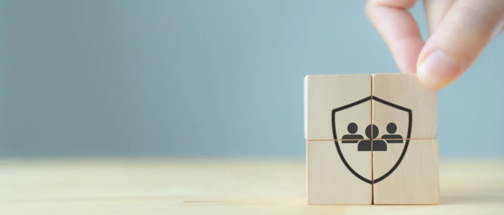 Close up of a hand placing wooden cube blocks together to form a shield icon enclosing a group of people symbol. The assembled blocks create a protective outline around the team figures against a soft blue background.