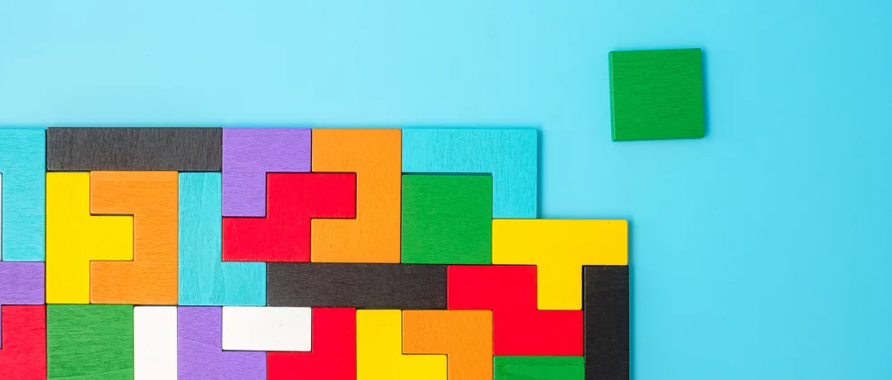 Colorful wooden puzzle blocks arranged tightly together on a bright blue background, forming an interlocking geometric pattern. A single green square block sits separated to the right, leaving a visible gap in the main arrangement.