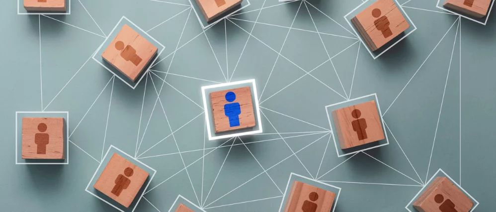 Overhead view of wooden cube blocks with person icons connected by white network lines across a muted background. One central block with a blue person icon is highlighted, standing out among surrounding brown icons.
