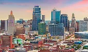 Downtown Kansas City, Missouri, at sunset with a colorful sky behind its mix of historic and modern skyscrapers. Streets below are lined with brick buildings, shops, and entertainment venues.