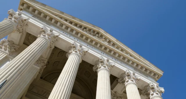 Low-angle view of a grand neoclassical building with tall Corinthian columns and ornate architectural details, set against a clear blue sky. The intricate carvings and symmetrical design convey authority and formality, often associated with government.