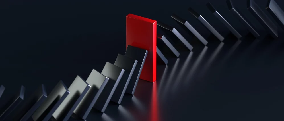 Row of black domino tiles falling in sequence across a dark reflective surface, halted by a single upright red domino tile. The red domino stands firmly in the center, preventing the chain reaction from continuing.