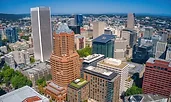 Aerial view of downtown Portland, Oregon, with a mix of modern skyscrapers and mid-rise buildings. The city is surrounded by greenery and distant hills under a clear blue sky.