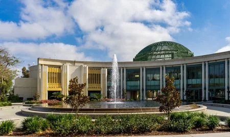 Elegant building with a green-domed roof and tall arched windows, centered behind a circular fountain spraying water upward. The scene is framed by manicured landscaping under a partly cloudy sky.