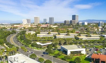 Aerial view of a business district with mid-rise office buildings surrounded by landscaped greenery and wide roads. Taller skyscrapers stand in the background beneath a bright blue sky with wispy clouds.