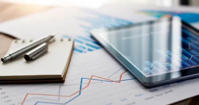 The image shows a close-up of a desk with a tablet, a pen, and a notepad placed on printed charts featuring line and bar graphs. The graphs display fluctuating data trends in red, blue, and gray, suggesting a focus on financial or business analysis.