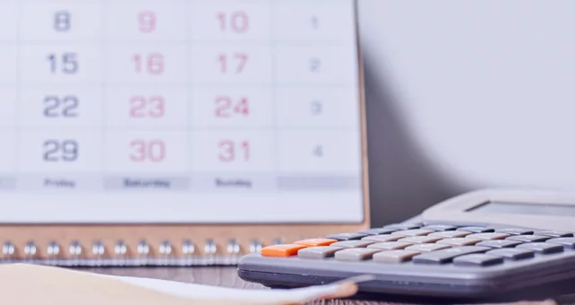 A desk scene shows a blurred monthly calendar in the background with a gray calculator and manila envelope in focus on a wooden surface. The setup suggests planning, scheduling, or financial calculations.