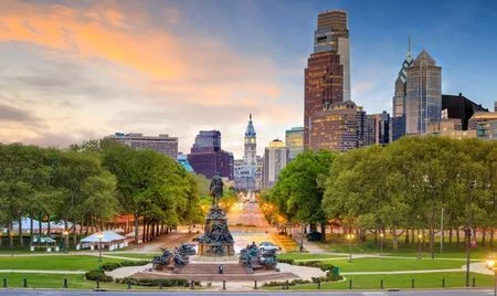 View of the Benjamin Franklin Parkway in Philadelphia, Pennsylvania, at sunset, with the Washington Monument Fountain in the foreground. The tree-lined boulevard leads toward City Hall, framed by the downtown skyline.