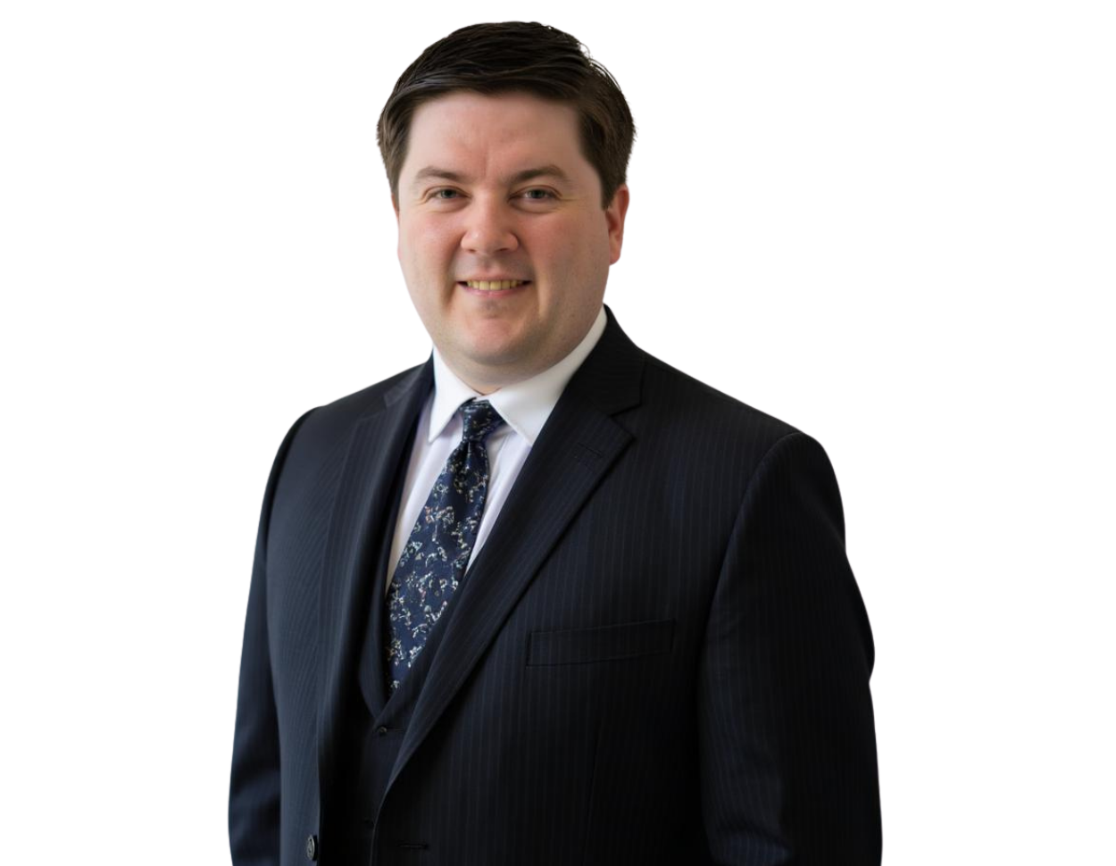 Smiling professional man with short dark hair wearing a dark pinstripe suit, white dress shirt, and patterned tie. He stands against a transparent background with a polished and approachable expression suited for a corporate or executive profile.
