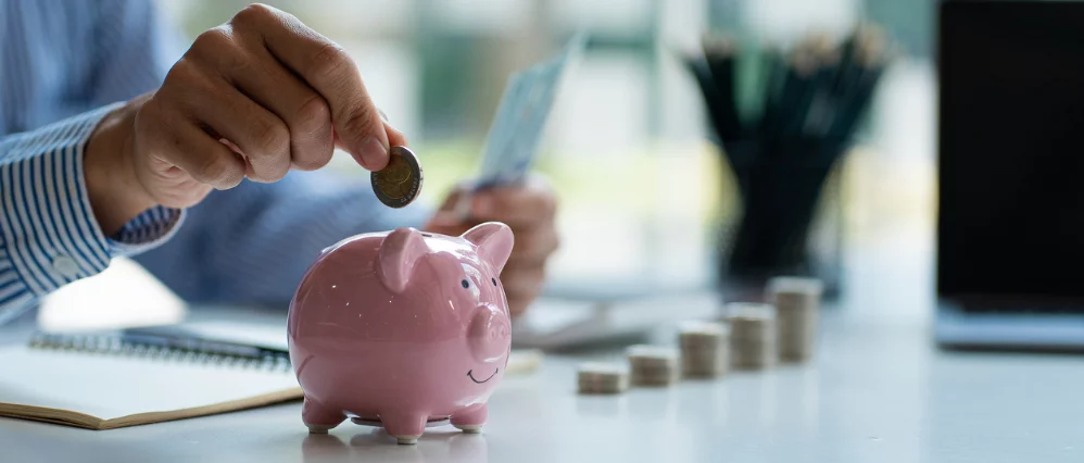 Close up of a person placing a coin into a pink ceramic piggy bank on a desk, while holding a credit card in the other hand. Stacks of coins are lined up in the background beside a notebook and laptop, suggesting budgeting or financial planning.