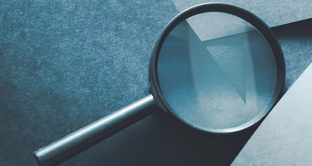 Close-up of a magnifying glass resting on textured blue paper with layered geometric shapes. The lens slightly distorts and enlarges the paper’s surface, creating a subtle play of light and shadow.