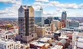 Aerial view of downtown Raleigh, North Carolina, with a mix of modern glass towers and historic brick buildings. The city stretches into the distance under a bright blue sky with scattered clouds.