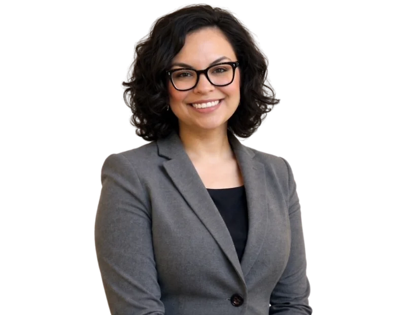 Smiling professional woman with short, dark curly hair and black-framed glasses wearing a gray blazer over a black top. She stands against a transparent background with a friendly, confident expression that conveys approachability and professionalism.