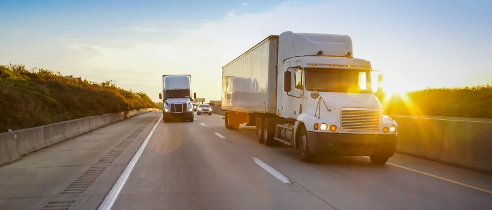 Semi tractor trailer trucks driving along a highway at sunset with warm golden light flaring across the scene. The lead white truck is in sharp focus while another truck and cars follow behind in the adjacent lane.
