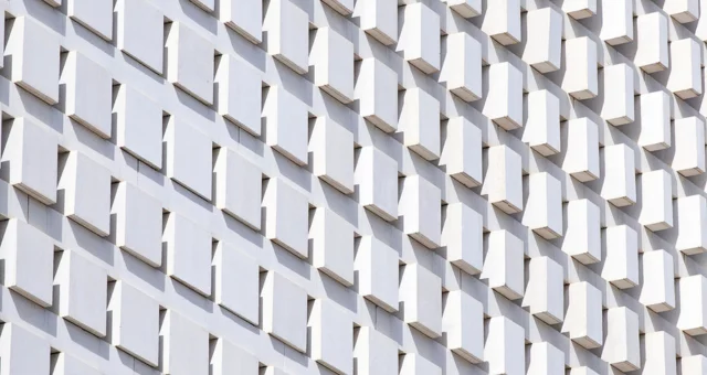 Facade of a modern building featuring a repetitive geometric pattern of protruding white rectangular blocks. The angled design creates shadows and depth, giving the surface a dynamic, textured appearance.