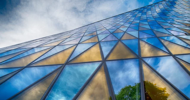 A modern glass building with triangular window panels reflects a vivid blue sky, white clouds, and green trees. The low-angle perspective emphasizes the geometric pattern as the structure appears to stretch infinitely upward.