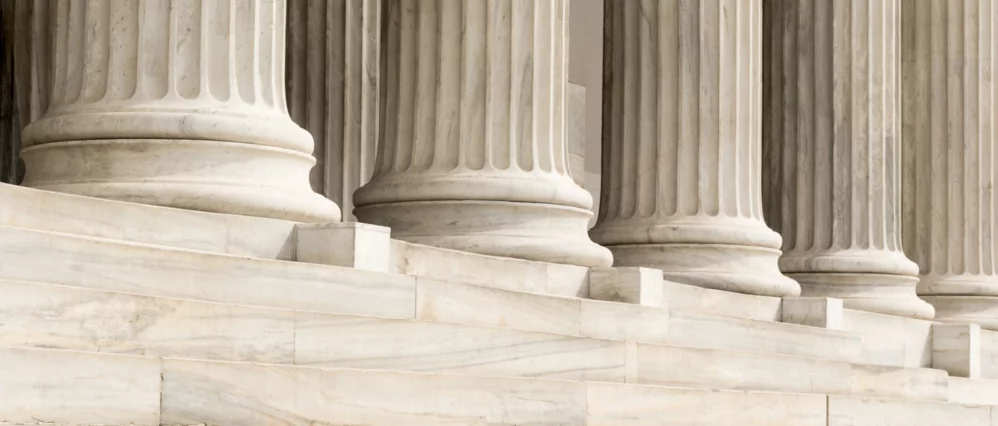 Close up of white marble steps leading up to a row of fluted Ionic columns with detailed bases and vertical grooves. The smooth stone surfaces and classical architectural lines emphasize symmetry and permanence.