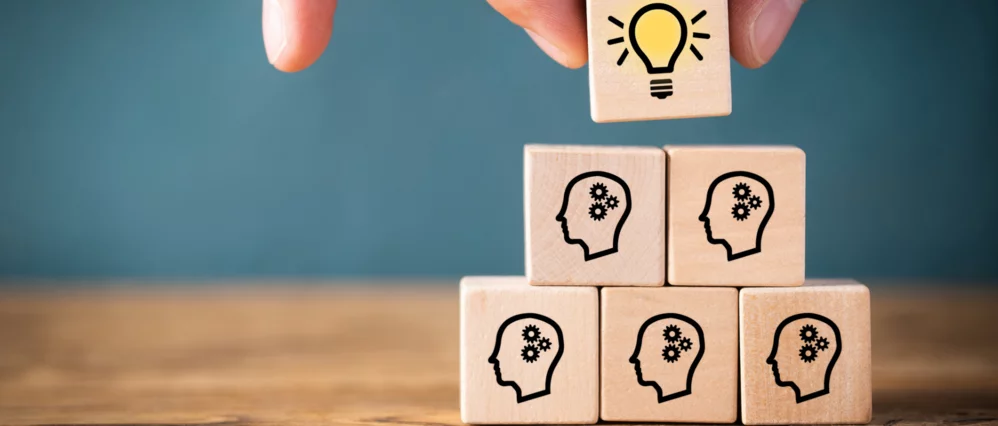 Close up of wooden cubes stacked in a pyramid on a table, each printed with a head silhouette containing gear icons. A hand places a top cube featuring a glowing light bulb symbol above the group.