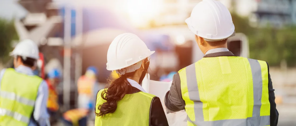Rear view of a businessman and businesswoman wearing white hard hats and high visibility safety vests reviewing construction plans at a job site. Other workers and equipment appear blurred in the background under warm sunlight.