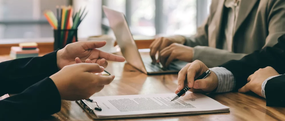 Close up of business professionals reviewing and signing a contract on a clipboard at a desk. One person points at the document while another holds a pen, with a laptop in the background.