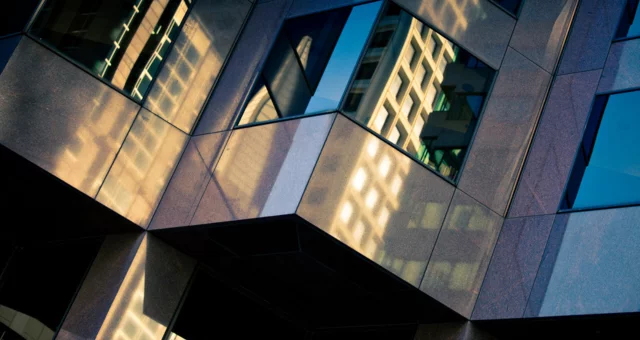 Close-up view of a modern glass and stone building facade reflecting distorted images of nearby structures. Warm sunlight enhances the golden tones of the reflections, contrasting with the cool blue of the sky in other window panels.