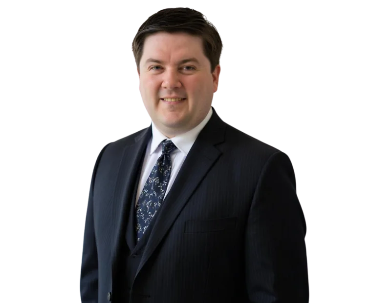 Smiling professional man with short dark hair wearing a dark pinstripe suit, white dress shirt, and patterned tie. He stands against a transparent background with a polished and approachable expression suited for a corporate or executive profile.