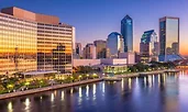 Evening view of downtown Jacksonville, Florida, with glass skyscrapers reflecting a vivid sunset. The St. Johns River runs along the foreground, its calm waters mirroring the city lights and colorful sky.
