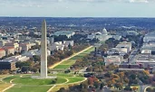 Aerial view of the National Mall in Washington, D.C., with the Washington Monument rising prominently in the foreground. The U.S. Capitol building is visible in the distance, surrounded by tree-lined streets and historic architecture.