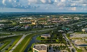 Aerial view of Port St. Lucie, Florida, showing a network of highways, commercial buildings, and residential neighborhoods. Green spaces and small waterways weave through the landscape under a partly cloudy sky.