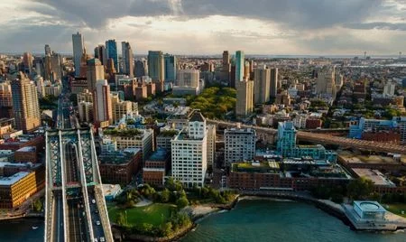 Aerial view of downtown Brooklyn, New York, with the Manhattan Bridge extending across the East River in the foreground. The skyline features a mix of modern high-rises and historic brick buildings surrounded by green spaces.