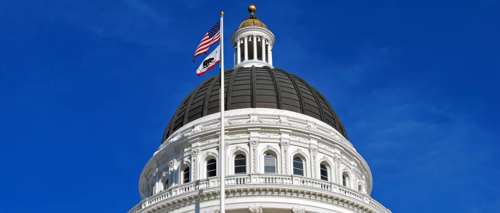 Capitol dome of the California State Capitol building in Sacramento with the United States flag and California state flag flying in front. The white neoclassical dome with ornate columns and a gold finial stands prominently against a clear deep blue sky.