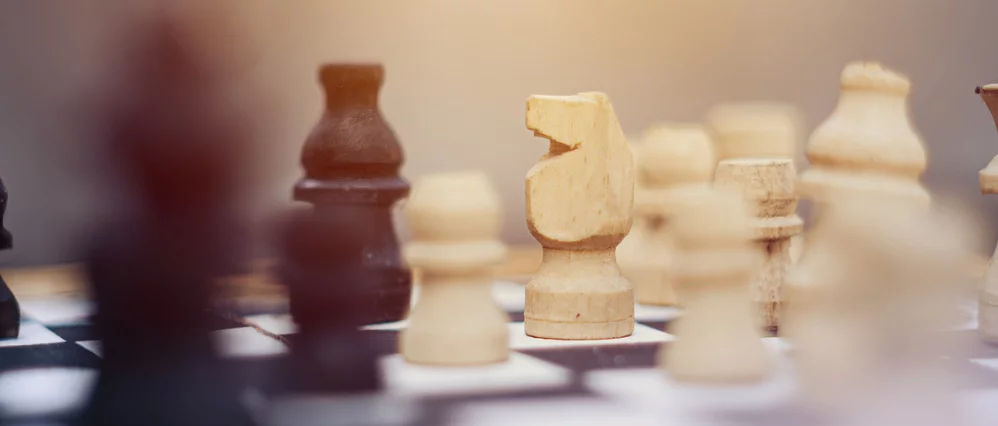 Close up of a wooden chessboard with a white knight piece in sharp focus at the center, surrounded by softly blurred chess pieces in the foreground and background. Warm sunset lighting casts a golden glow across the board, creating a shallow depth of fiel