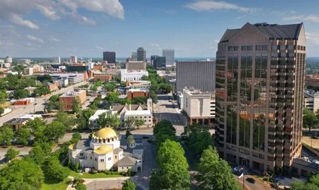 Aerial view of downtown Columbia, South Carolina, with modern office towers and tree-lined streets. The gold-domed building of the St. Peter’s Catholic Church stands out in the foreground.