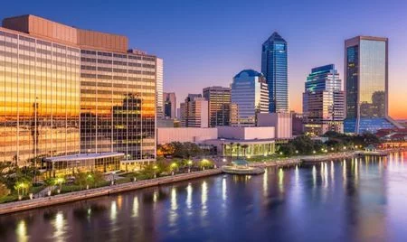 Evening view of downtown Jacksonville, Florida, with glass skyscrapers reflecting a vivid sunset. The St. Johns River runs along the foreground, its calm waters mirroring the city lights and colorful sky.