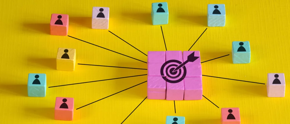 Overhead view of colorful cube blocks with person icons arranged around a central cluster of pink cubes featuring a target symbol with an arrow in the bullseye. Black lines connect each surrounding block to the center, forming a clear network pattern on a