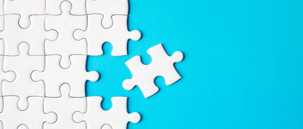 Directly above view of white jigsaw puzzle pieces arranged on a bright blue background, with one loose piece separated to the right. The connected pieces form a partial grid, leaving a visible gap where the missing piece would fit.
