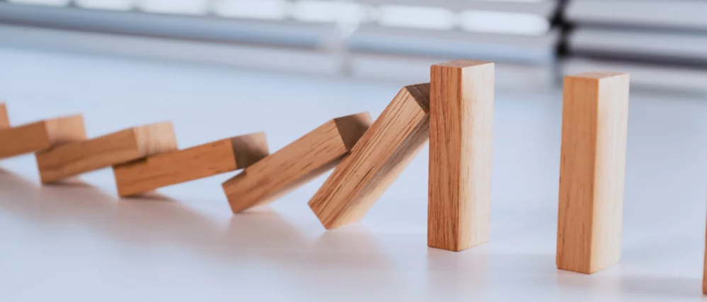 Close up of wooden domino blocks falling in sequence on a white surface, stopped by a thicker upright wooden block at the end. The stronger vertical block prevents the chain reaction from continuing, creating a clear break in the line.