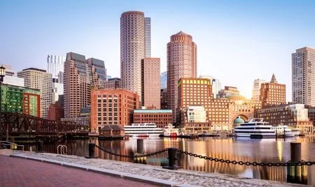 Boston’s waterfront skyline in early morning light, with modern and historic brick buildings reflecting in the calm harbor. Docked boats and a chain-lined walkway frame the foreground.