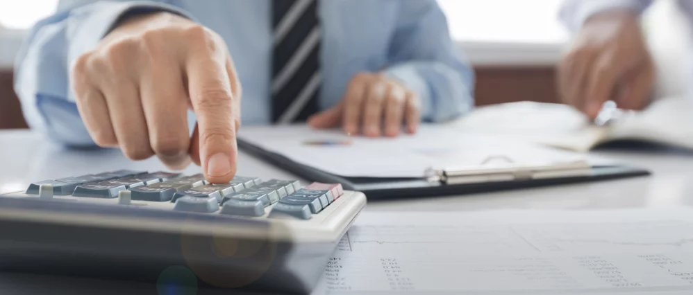 Close up of a business professional pressing keys on a calculator while reviewing printed financial documents on a desk. A clipboard with reports and charts sits nearby, suggesting detailed accounting or budgeting work.