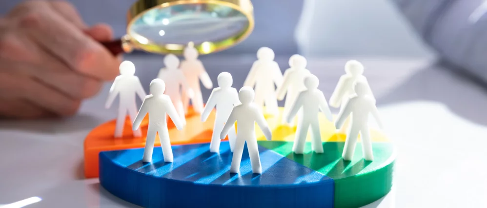 Close up of white human figure icons standing on a colorful segmented pie chart while a hand holds a magnifying glass above them. The lens focuses on several figures, suggesting evaluation or selection within a group.