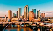 Downtown Tampa, Florida, at golden hour with sunlight reflecting off its tall glass and brick skyscrapers. A bridge crosses the Hillsborough River in the foreground, connecting the urban core with surrounding neighborhoods.