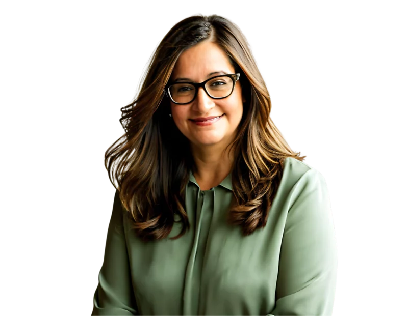 A woman with long, wavy brown hair and glasses smiles softly while wearing a sage green blouse. She is posed confidently with her hands loosely clasped, suggesting a warm and approachable demeanor.