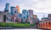 Minneapolis skyline with modern skyscrapers reflecting the evening light. The historic Stone Arch Bridge spans the Mississippi River in the foreground, leading toward the city.