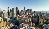 A bright daytime view of a modern city skyline with tall glass and steel skyscrapers rising above dense mid-rise buildings. Mountains are visible in the background under a clear blue sky.