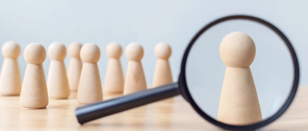 Close up of wooden peg figures arranged in a row on a table, with one figure enlarged through a magnifying glass in the foreground. The focused figure stands out sharply while others remain blurred in the background.
