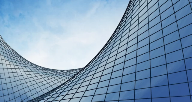 Curved glass facade of a modern skyscraper reflecting the blue sky, creating a flowing, wave-like architectural form. The sleek grid of windows emphasizes both elegance and futuristic design.