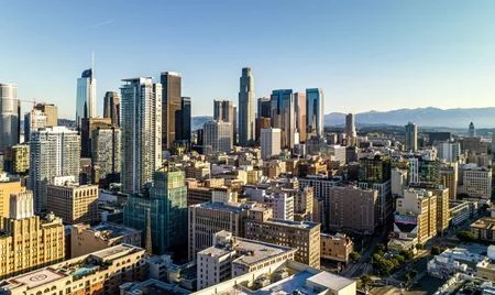 A bright daytime view of a modern city skyline with tall glass and steel skyscrapers rising above dense mid-rise buildings. Mountains are visible in the background under a clear blue sky.